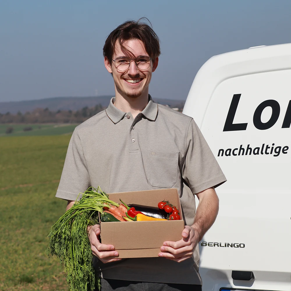Joshua von Ameln von LomoGro aus Düren hält einen Gemüsekorb in der Hand und steht auf einer Wiese vor seinem Elektro-Transporter.